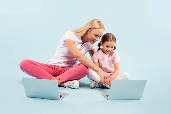 Sorridente madre e figlia seduta con le gambe incrociate vicino ai computer portatili su blu — Foto stock