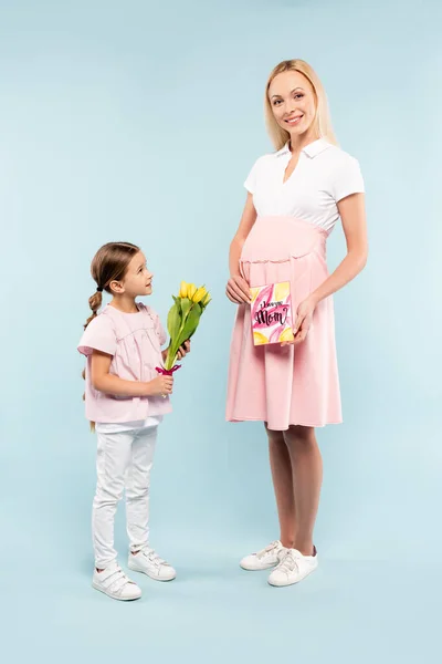 Полный рост малыша, держащего тюльпаны рядом со счастливой беременной мамой на голубом фоне — стоковое фото