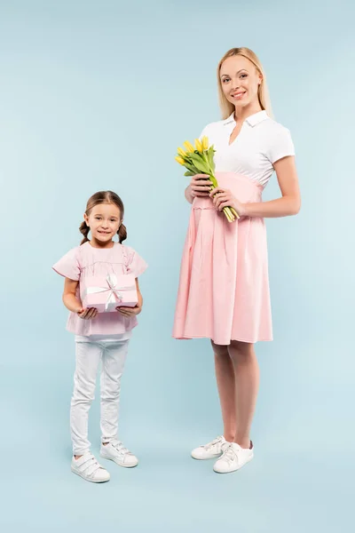 Longitud completa de niño sosteniendo presente cerca de la madre embarazada con tulipanes en azul - foto de stock