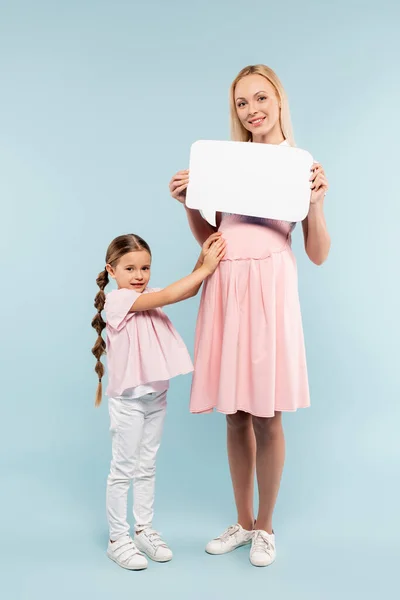 Full length of pregnant mother holding speech bubble near kid on blue — Stock Photo