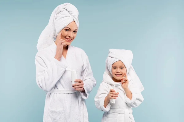 Mãe feliz e criança em roupões de banho segurando tubos e aplicando creme cosmético em rostos isolados em azul — Fotografia de Stock