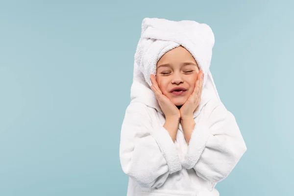 Niño feliz en albornoz y toalla en la cabeza sonriendo mientras toca las mejillas aisladas en azul - foto de stock