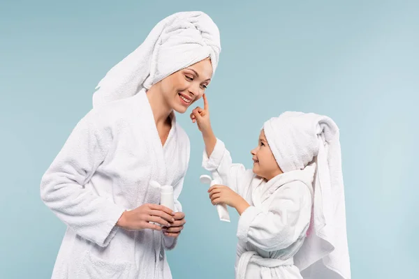 Happy kid in bathrobe holding tube while applying cosmetic cream on face of mother isolated on blue — Stock Photo