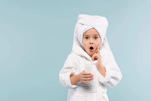 Shocked kid in bathrobe and towel on head holding tube with cosmetic product isolated on blue — Stock Photo
