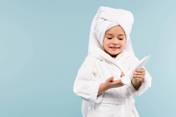 Улыбающийся ребенок в халате и полотенце на головной упор трубки с косметическим продуктом изолированы на синий — стоковое фото
