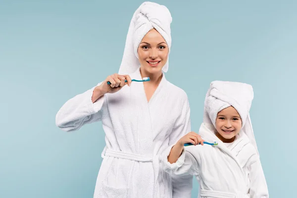 Sonriente madre y niño en albornoces sosteniendo cepillos de dientes con pasta de dientes aislados en azul - foto de stock