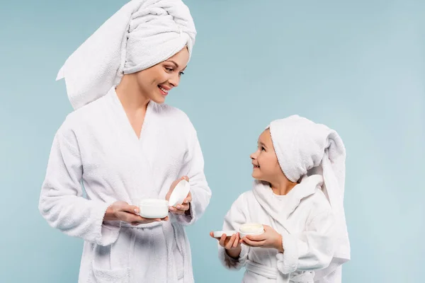 Smiling mother and kid in bathrobes holding containers with cosmetic cream isolated on blue — Stock Photo