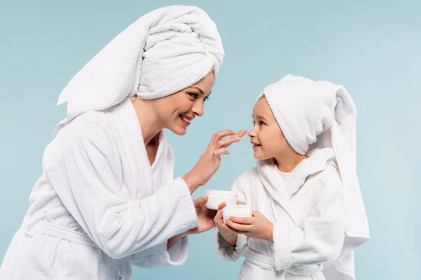 Madre felice in accappatoio che tiene il contenitore e applica la crema cosmetica sul viso della figlia isolata sul blu — Foto stock