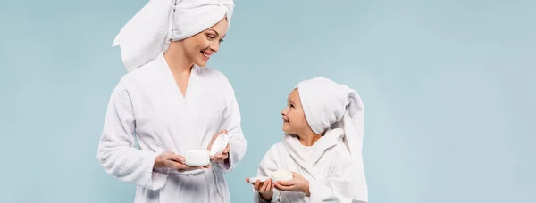 Heureux mère et enfant dans des peignoirs contenant des récipients avec crème cosmétique isolé sur bleu, bannière — Photo de stock