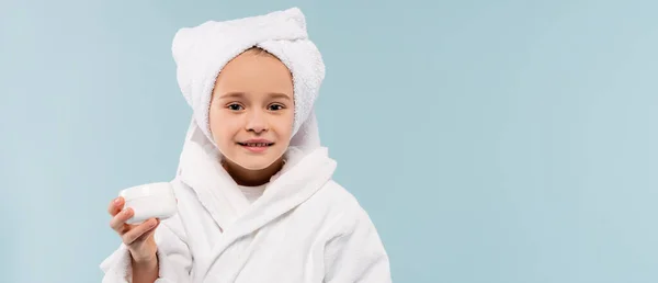 Criança sorridente em roupão de banho e toalha na cabeça segurando recipiente com produto cosmético isolado em azul, banner — Fotografia de Stock