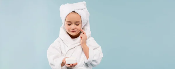 Улыбающийся ребенок в купальном халате, держащий контейнер и наносящий косметический продукт, изолированный на синий, баннер — стоковое фото