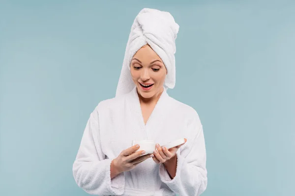 Amazed woman in bathrobe holding container with cosmetic cream isolated on blue — Stock Photo