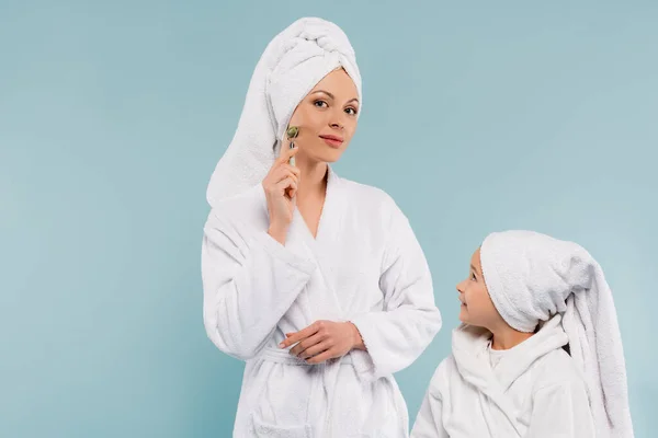 Happy kid in bathrobe looking at mother using jade roller isolated on blue — Stock Photo