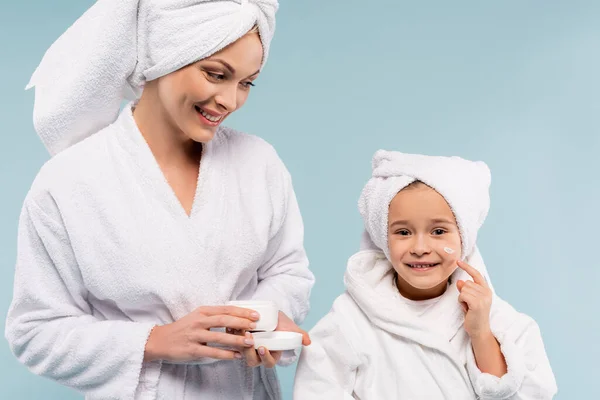 Mãe feliz em roupão segurando recipiente e olhando para a filha aplicando creme cosmético no rosto isolado no azul — Fotografia de Stock