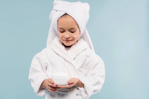 Fröhliches Kind in Bademantel und Handtuch auf dem Kopf beim Betrachten eines Behälters mit einem auf blau isolierten Kosmetikprodukt — Stockfoto