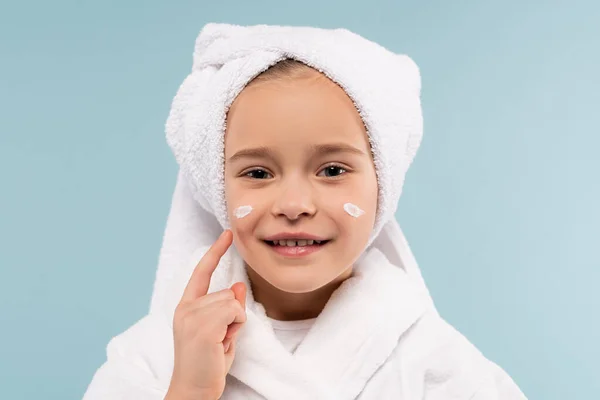 Fröhliches Kind in Bademantel und Handtuch auf dem Kopf, kosmetische Creme isoliert auf blau auftragen — Stockfoto