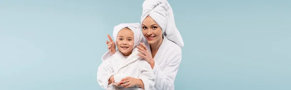 Glückliche Mutter im Bademantel, die kosmetische Creme auf das Gesicht ihrer Tochter aufträgt, isoliert auf blauem Grund, Banner — Stockfoto