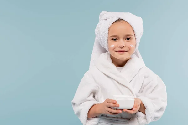 Cheerful child in bathrobe and cosmetic cream on face holding container isolated on blue — Stock Photo