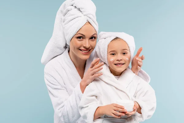 Feliz madre en albornoz aplicando crema cosmética en la cara de la hija aislada en azul - foto de stock