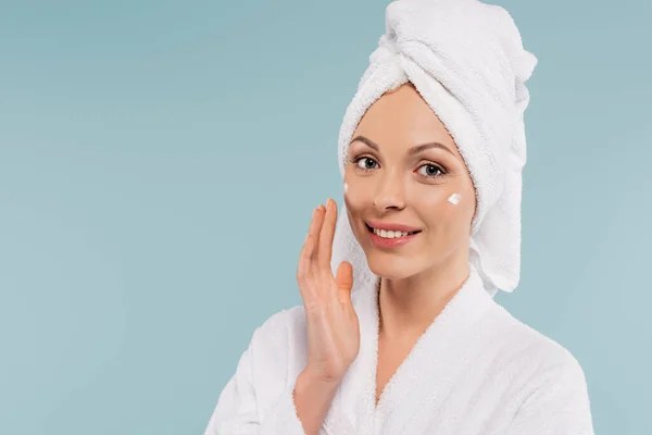 Mujer sonriente en albornoz blanco aplicando crema facial aislada en azul - foto de stock