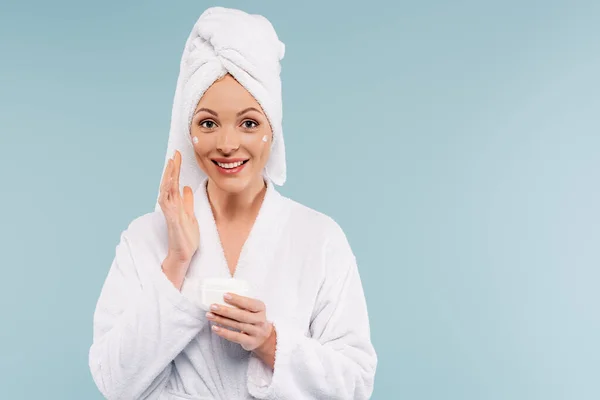 Mulher feliz em roupão de banho branco segurando recipiente com creme facial isolado em azul — Fotografia de Stock