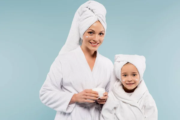 Happy mother in bathrobe holding container with face cream near daughter isolated on blue — Stock Photo