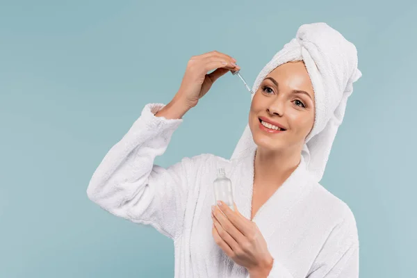 Happy mother in bathrobe holding bottle and applying serum isolated on blue — Stock Photo