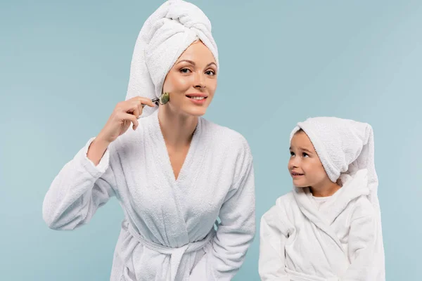 Niño alegre en albornoz mirando madre feliz usando rodillo de jade aislado en azul - foto de stock