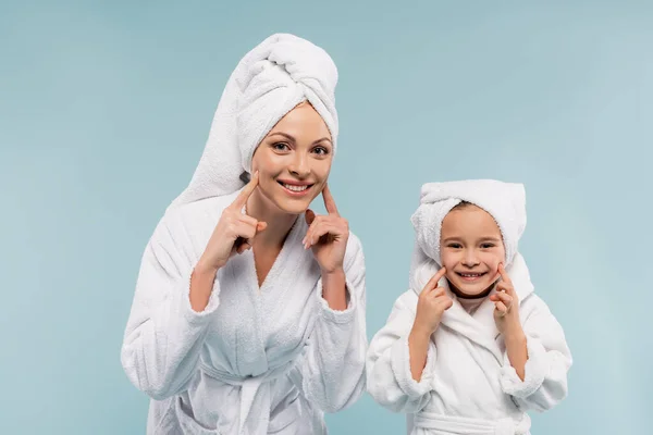 Mãe alegre e criança em roupões de banho sorrindo e tocando bochechas isoladas em azul — Fotografia de Stock