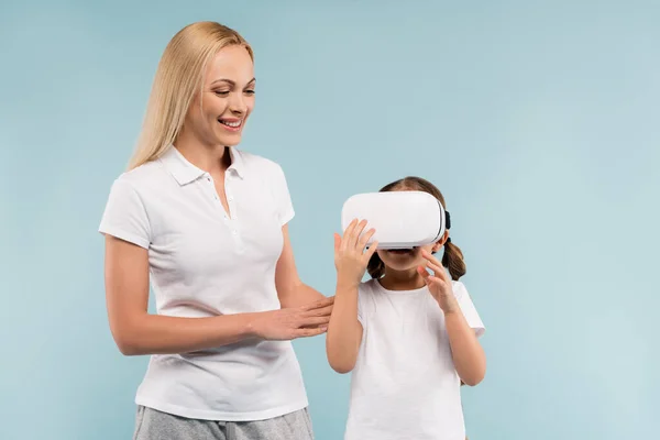 Веселая мать смотрит на дочь в VR гарнитуре изолированы на синий — стоковое фото