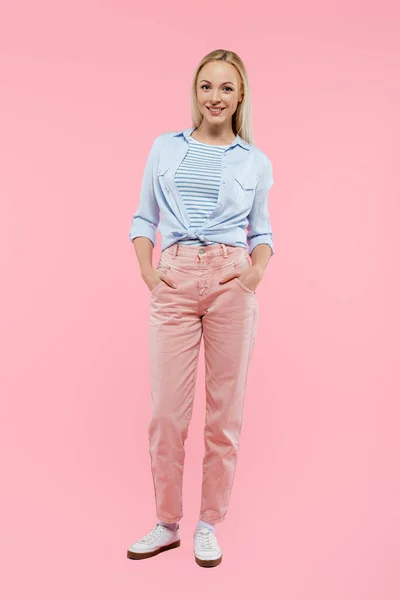 Full length of happy and blonde woman smiling while standing with hands in pockets on pink — Stock Photo