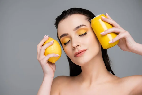 Брюнетка молода жінка з закритими очима тримає жовті банки з безалкогольним напоєм і лимоном ізольовані на сірому — стокове фото