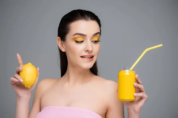 Brünette junge Frau hält gelbe Dose mit Erfrischungsgetränk und Zitrone und beißt Lippen isoliert auf grau — Stockfoto