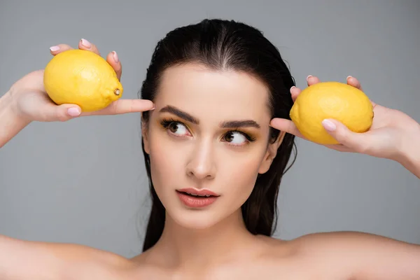 Brunette woman with bare shoulders holding yellow and ripe lemons isolated on grey — Stock Photo
