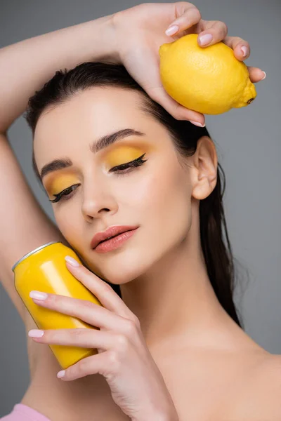 Brünette junge Frau mit gelber Dose mit Softdrink und Bio-Zitrone isoliert auf grau — Stockfoto