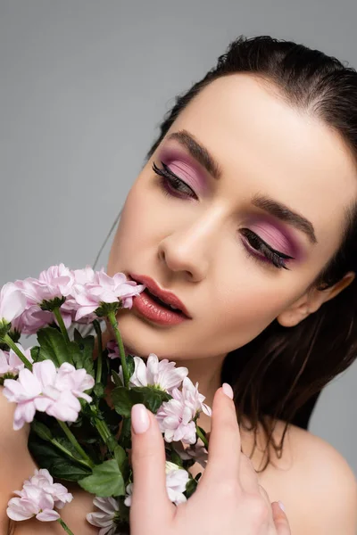 Mujer joven con sombras de ojos rosados mirando hacia otro lado cerca de flores aisladas en gris - foto de stock