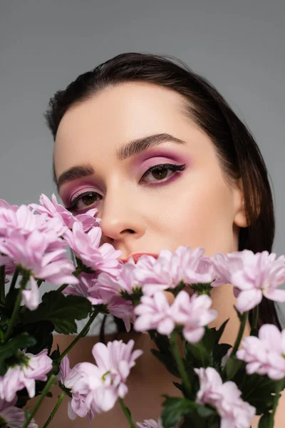 Jeune femme avec des ombres à paupières roses regardant la caméra à travers des fleurs isolées sur gris — Photo de stock