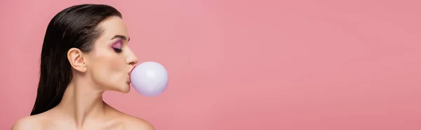 Side view of brunette woman with bare shoulders blowing bubblegum isolated on pink, banner — Stock Photo