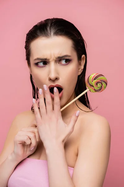 Shocked young woman covering mouth and holding lollipop isolated on pink — Stock Photo