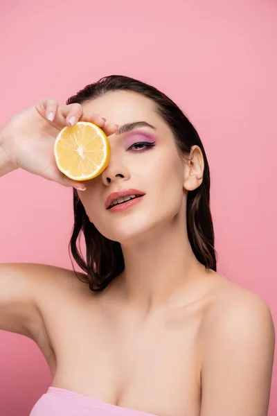 Mujer joven con los hombros desnudos cubriendo el ojo con la mitad de limón maduro y mirando a la cámara aislada en rosa - foto de stock