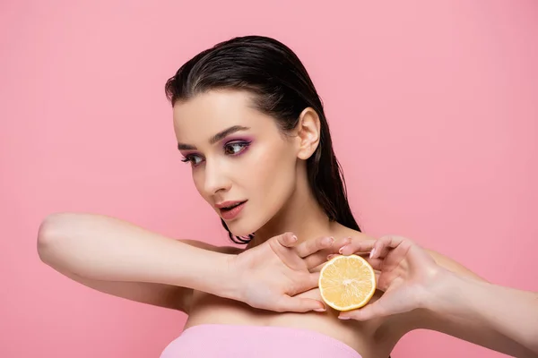 Mujer joven con hombros desnudos sosteniendo la mitad de limón maduro y mirando hacia otro lado aislado en rosa - foto de stock