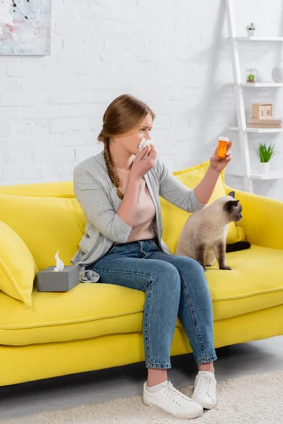 Donna con allergia guardando vaso con pillole vicino gatto sul divano giallo a casa — Foto stock