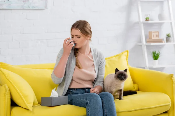 Mulher com alergia usando inalador perto de gato siamês peludo no sofá — Fotografia de Stock