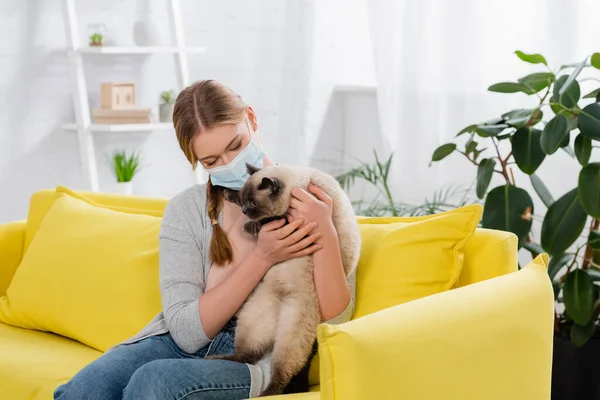 Mujer joven con reacción alérgica usando máscara médica y sosteniendo gato peludo en un sofá amarillo - foto de stock