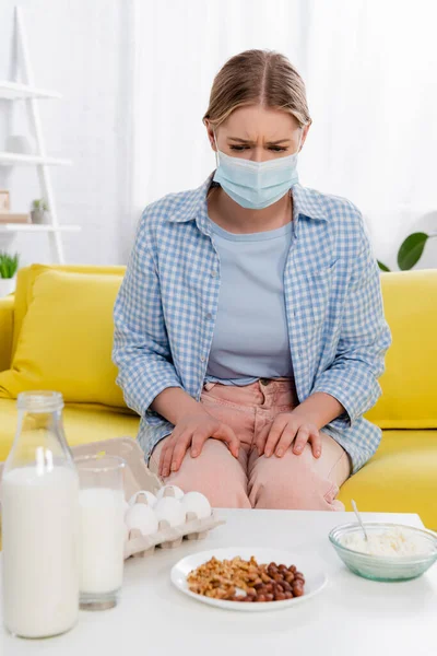 Woman with allergy wearing medical mask near milk, cottage cheese and eggs on blurred foreground - foto de stock