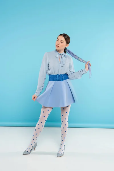 Mujer de moda mirando hacia otro lado y tocando el pañuelo mientras posa sobre fondo azul - foto de stock