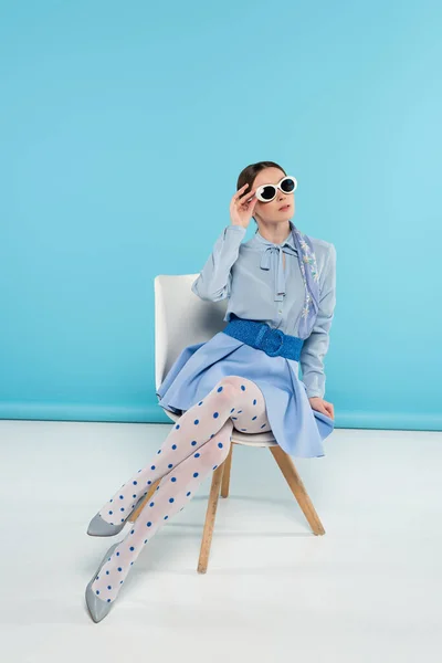 Stylish woman adjusting sunglasses while sitting on blue background — Stock Photo