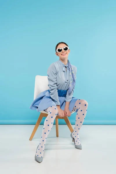 Smiling trendy woman in dotted tights and sunglasses sitting on blue background — Stock Photo