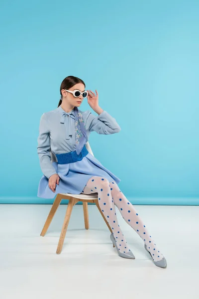 Mujer extravagante tocando gafas de sol mientras está sentado sobre fondo azul - foto de stock