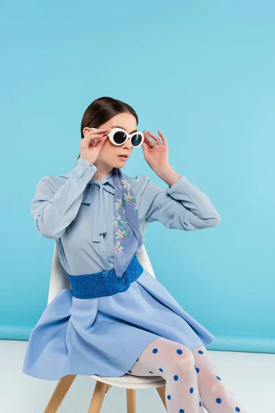 Elegant woman adjusting sunglasses while sitting on blue background — Stock Photo